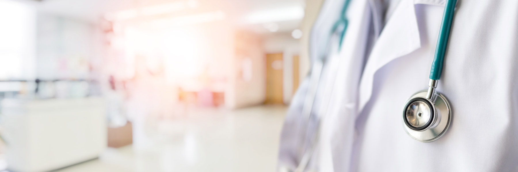 Close-up of a healthcare professional wearing a white coat and stethoscope, standing in a bright clinical setting with a softly blurred hospital interior in the background.