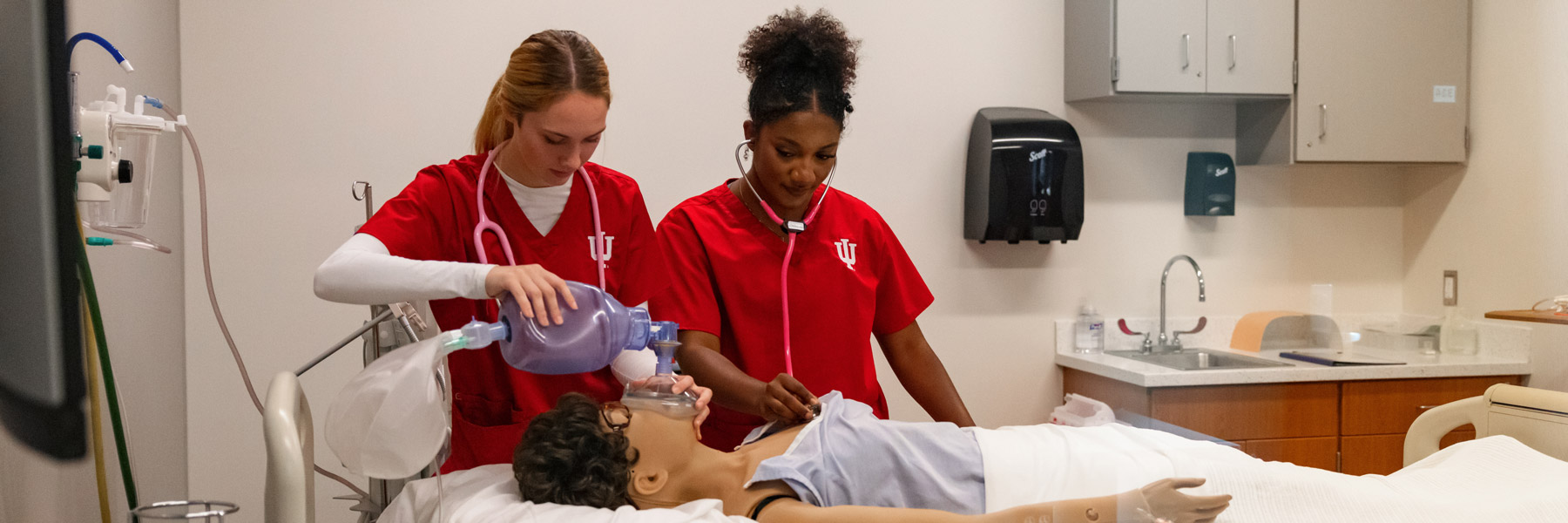 Indiana University School of Nursing students in red scrubs practicing airway management on a simulation mannequin in a clinical lab setting.
