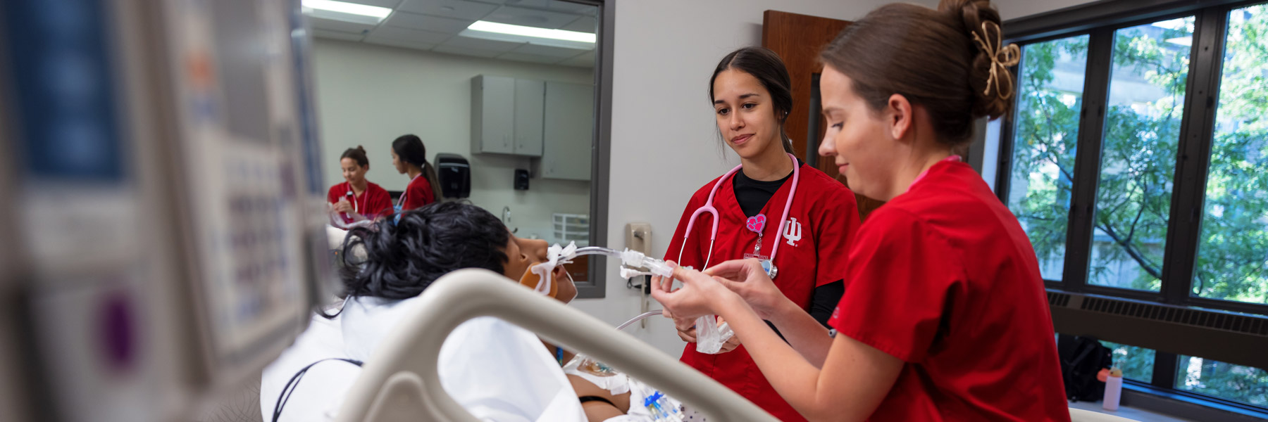IU School of Nursing students in red scrubs provide care to a patient manikin in a simulated hospital room.
