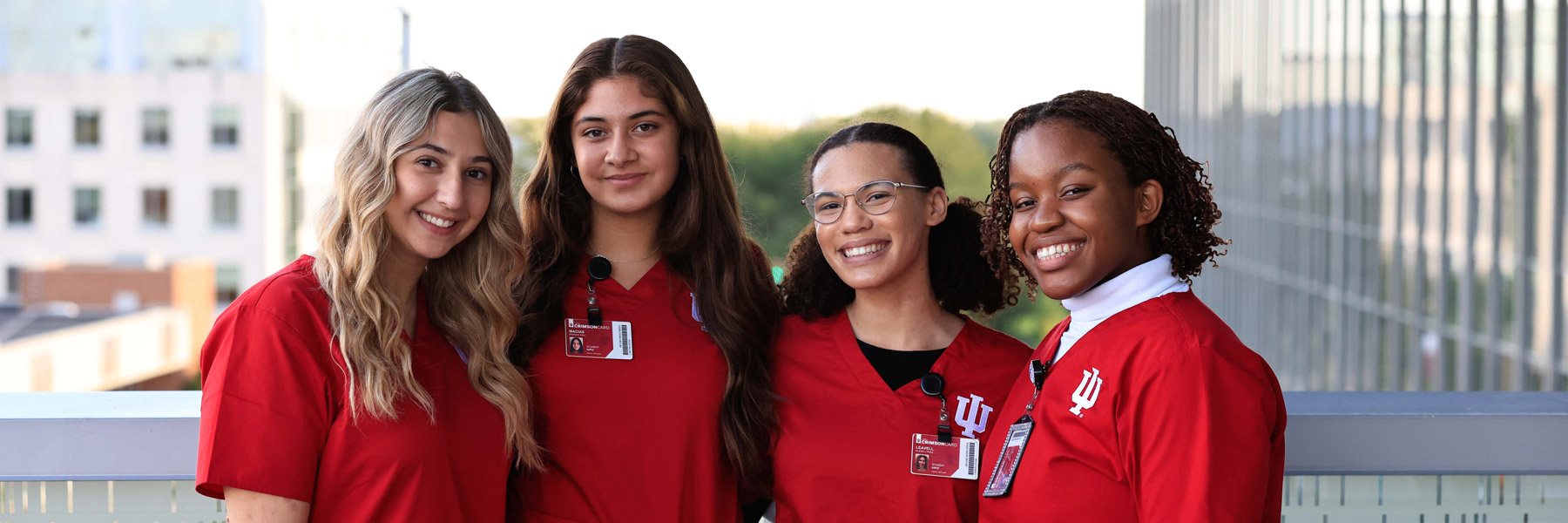 Four Indiana University School of Nursing students in red scrubs stand together on an outdoor campus balcony, smiling with their arms around one another.