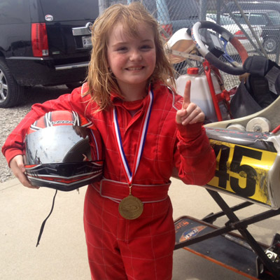 A young Maxine Cosat in a red racing suit smiles while holding a helmet and wearing a medal, standing beside a go-kart and raising one finger to signal first place.