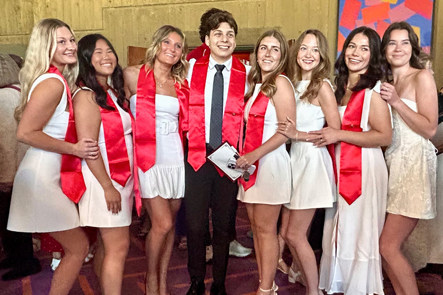 A group of graduates dressed in white outfits and red stoles gather together and smile for a celebratory photo at a commencement event.