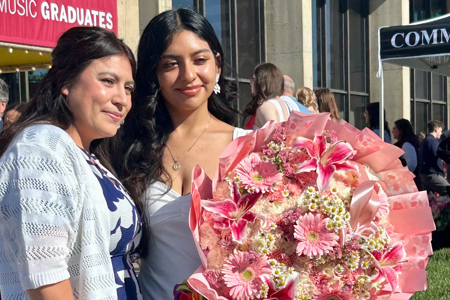 Two women stand together outside after a graduation ceremony, smiling beside a large bouquet of pink flowers in bright sunlight.