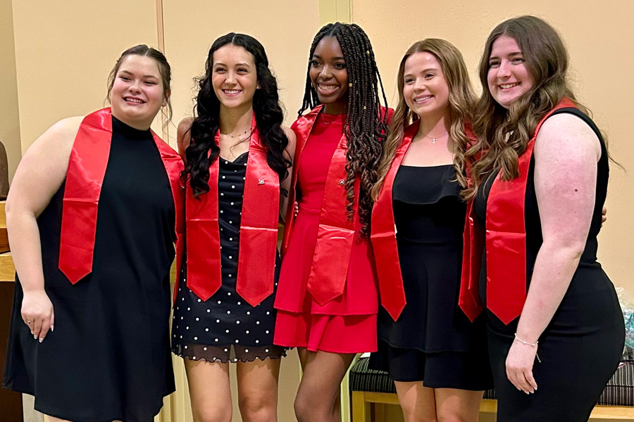 Five graduates wearing red stoles pose together and smile for a photo during a commencement celebration.