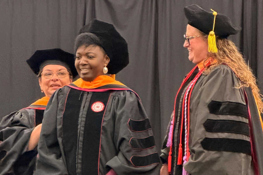 A graduate is hooded by two faculty during a commencement ceremony, smiling and celebrating together.
