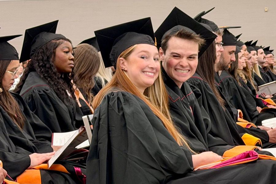 Graduating students in caps and gowns sit together during a commencement ceremony, with two smiling graduates looking toward the camera in the foreground.