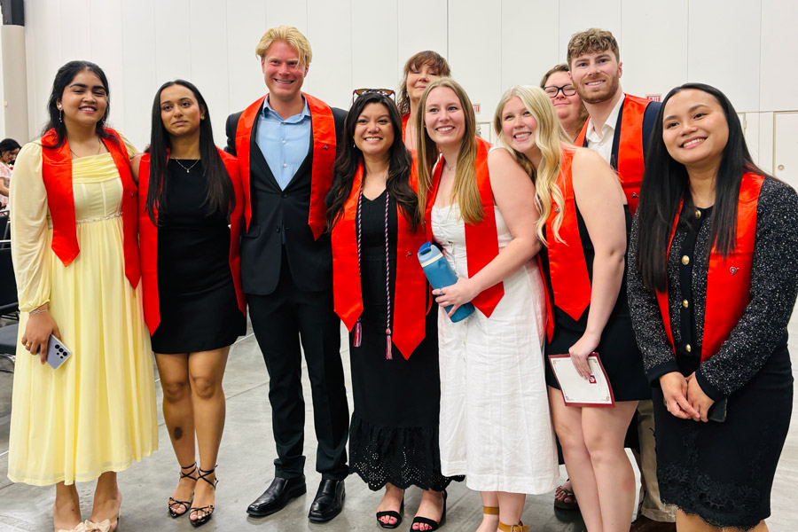 A group of graduates wearing red stoles pose together and smile for a photo during a commencement celebration.