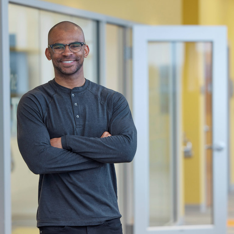 Jordan Page poses for a photo with his arms crossed while standing outside of an open classroom door