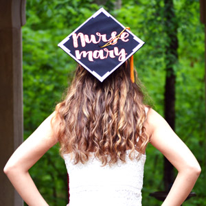 A girl with her back turned toward the camera wears a graduation cap with "Nurse Mary" on it.