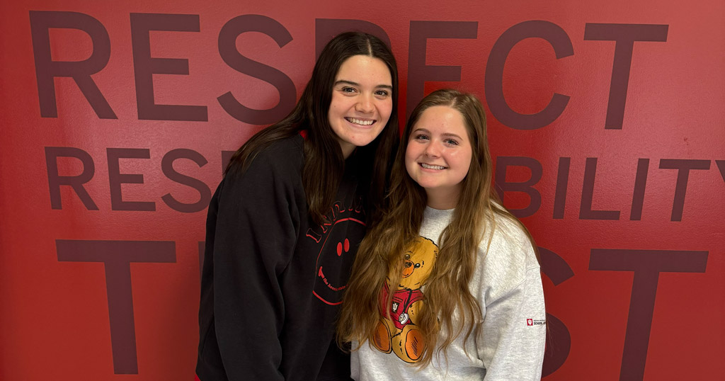 Kara Bennett and Paige Collins stand smiling in front of a red wall displaying the words, Respect, Responsibility, Trust.