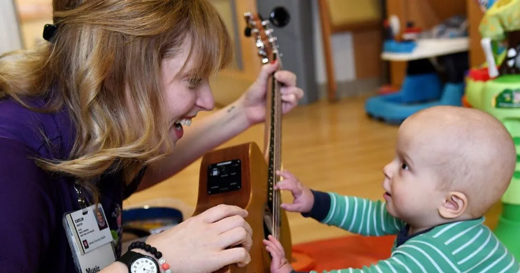 A music therapist smiles while holding a guitar as an infant reaches out to touch it during a therapy session.