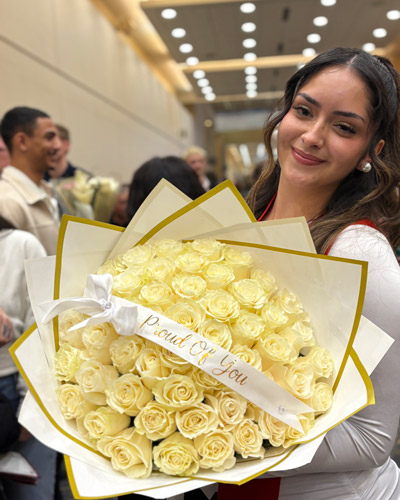 A December 2025 graduate holds a large flower bouquet with a ribbon that states Proud of You.