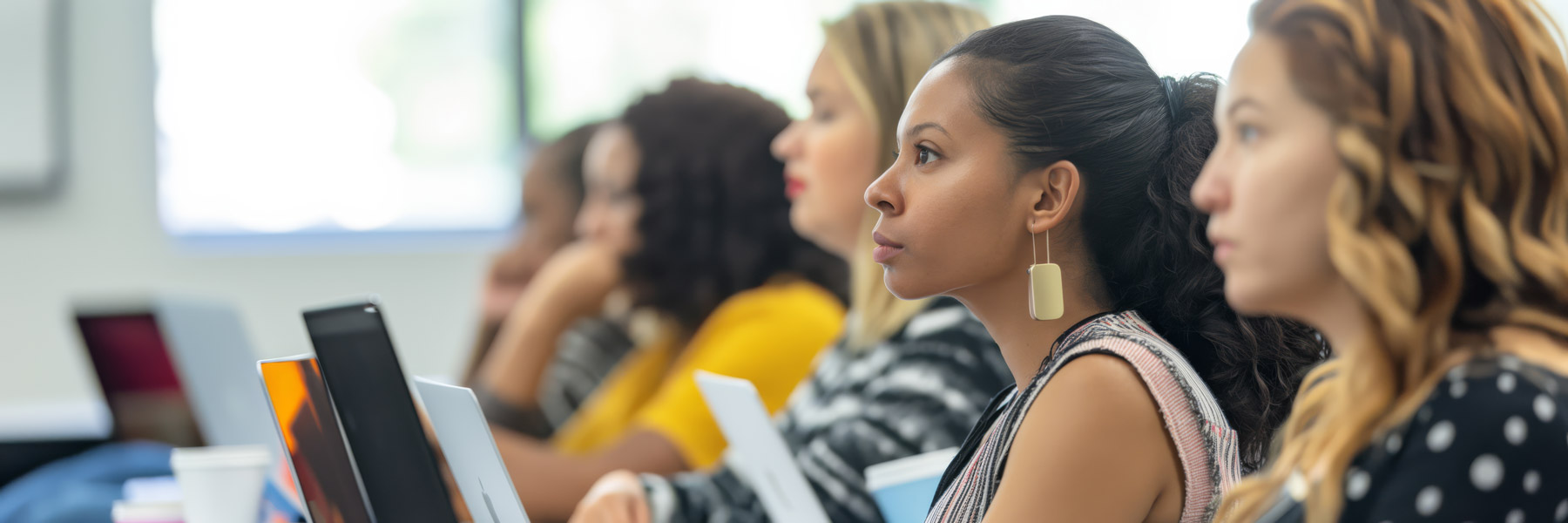 Group of adult students sitting in a classroom, focused on a lecture with laptops open.
